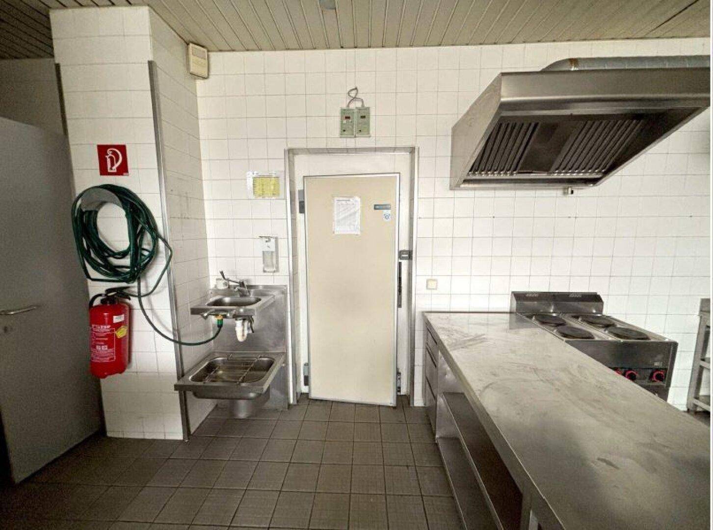 Professionelle Großküche mit Edelstahlmöbeln, Fliesenwänden und einer großen Dunstabzugshaube.