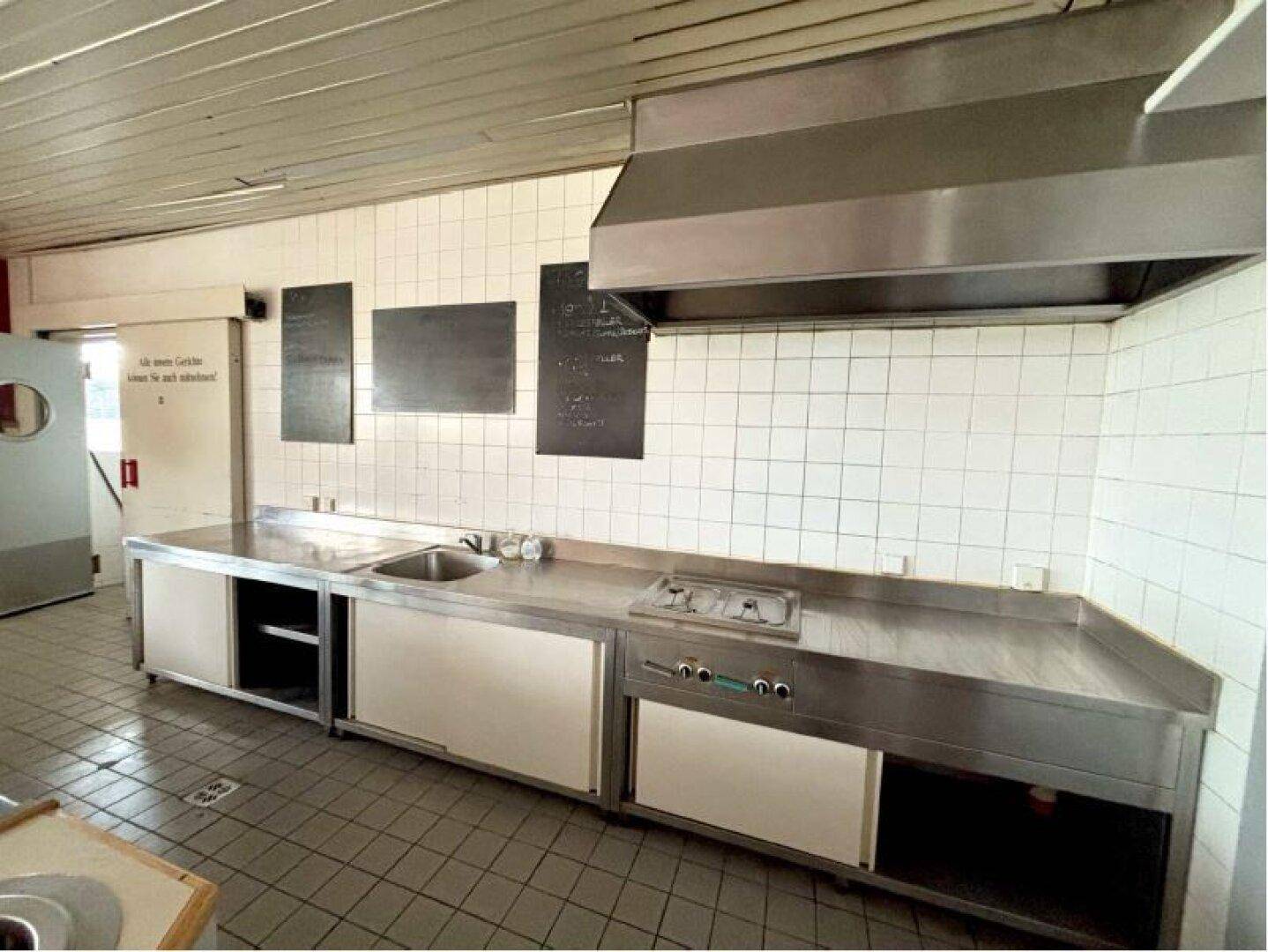 Arbeitsbereich einer Großküche mit Edelstahltheke, Spülbecken, Kochfeld und Dunstabzugshaube.