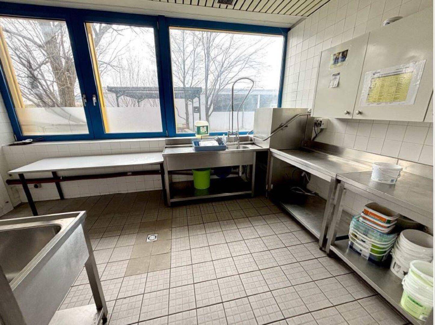 Spülbereich einer Großküche mit großen Fenstern, Edelstahlspülbecken und gefliesten Wänden.