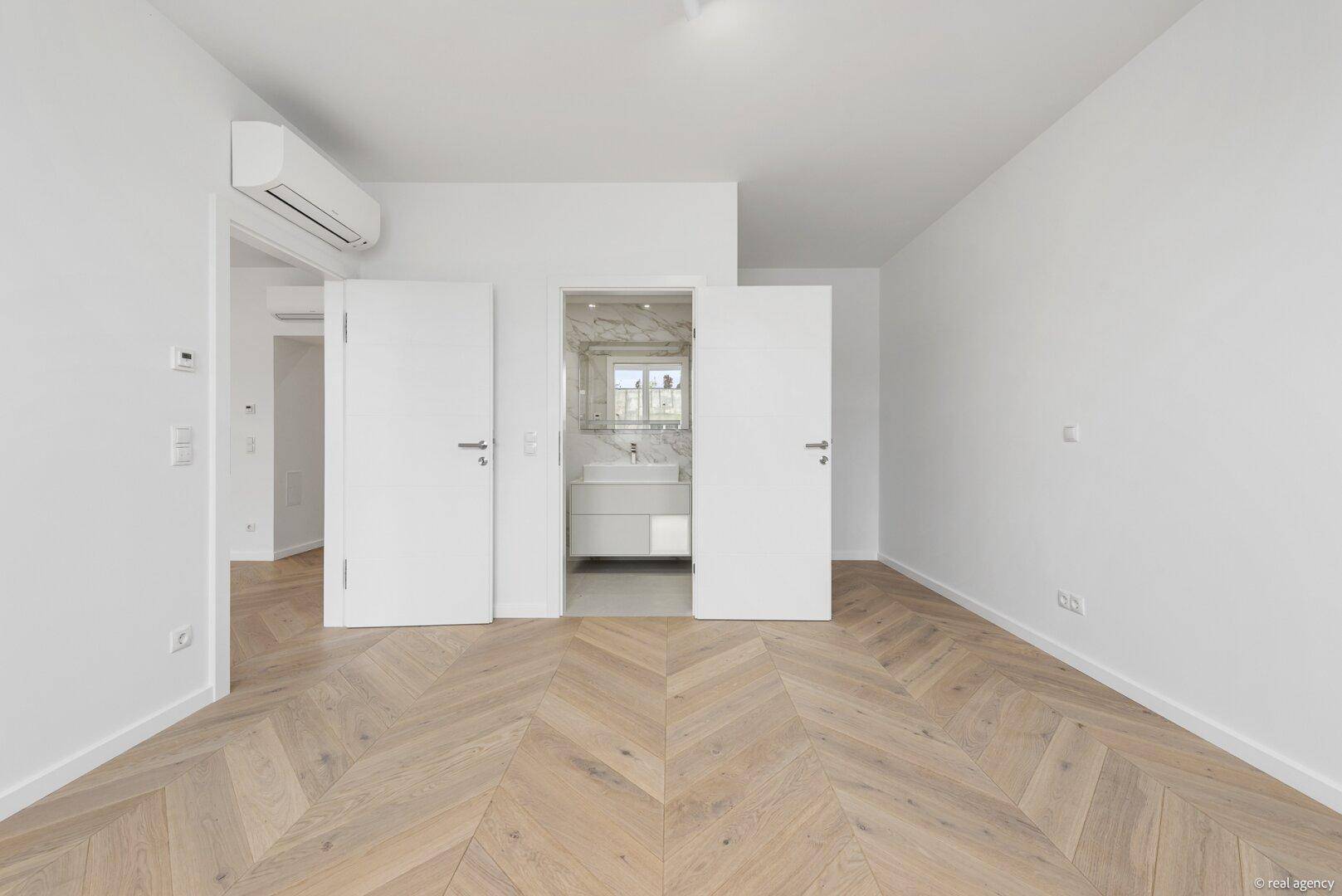 Schlafzimmer mit Fischgrätparkett und direktem Zugang zu einem modernen Badezimmer mit Marmorfliesen.