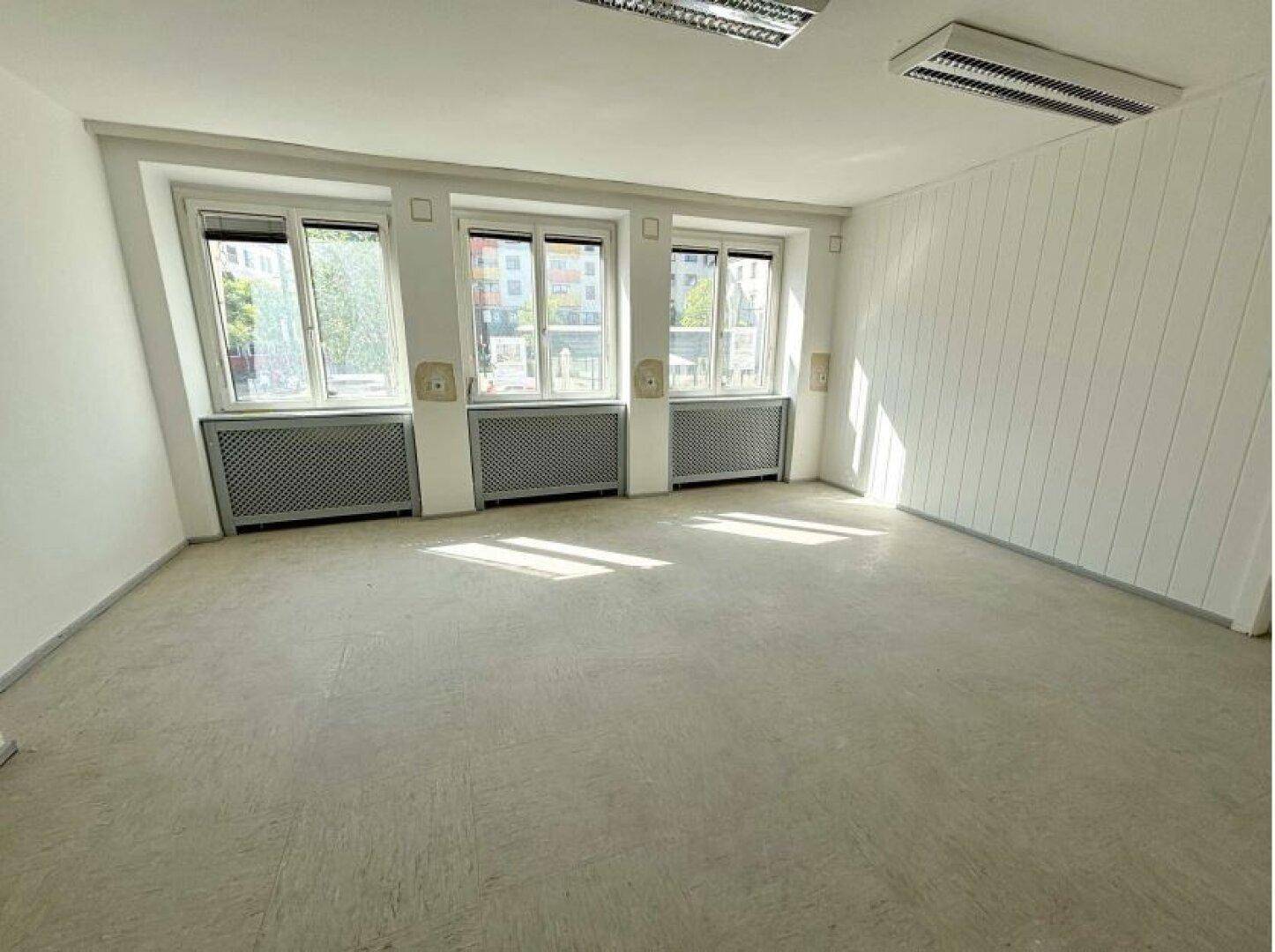 Ein heller, leerer Büroraum mit drei großen Fenstern, die viel Tageslicht hereinlassen, und Heizkörpern darunter.