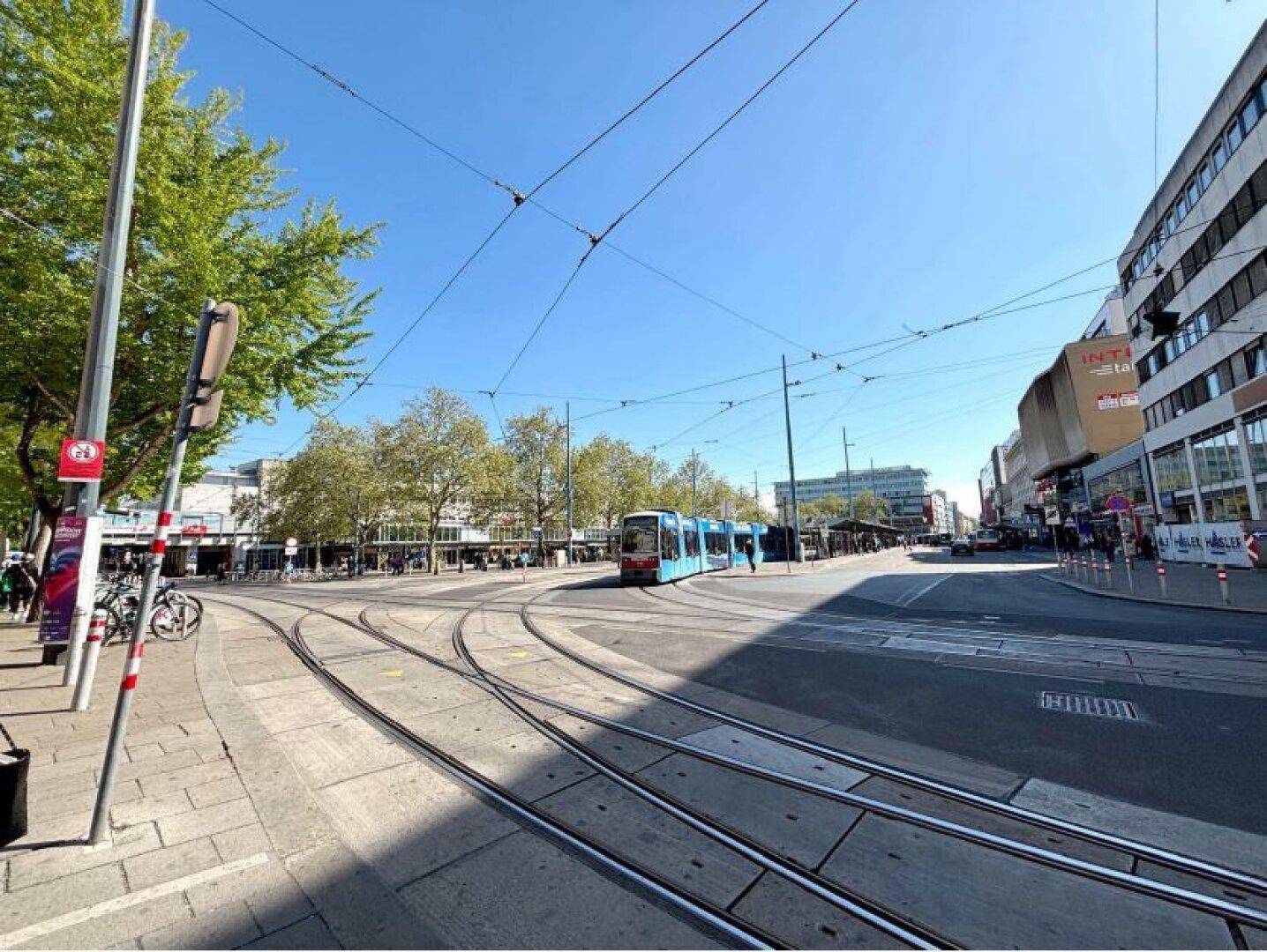 Eine belebte Stadtstraße mit Straßenbahnschienen, Gebäuden und Bäumen unter einem klaren blauen Himmel.
