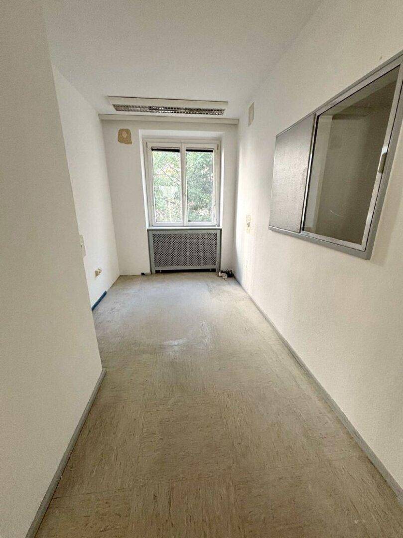 Ein schmaler Büroraum oder Flur mit einem Fenster, das Tageslicht hereinlässt, und einem funktionalen Bodenbelag.
