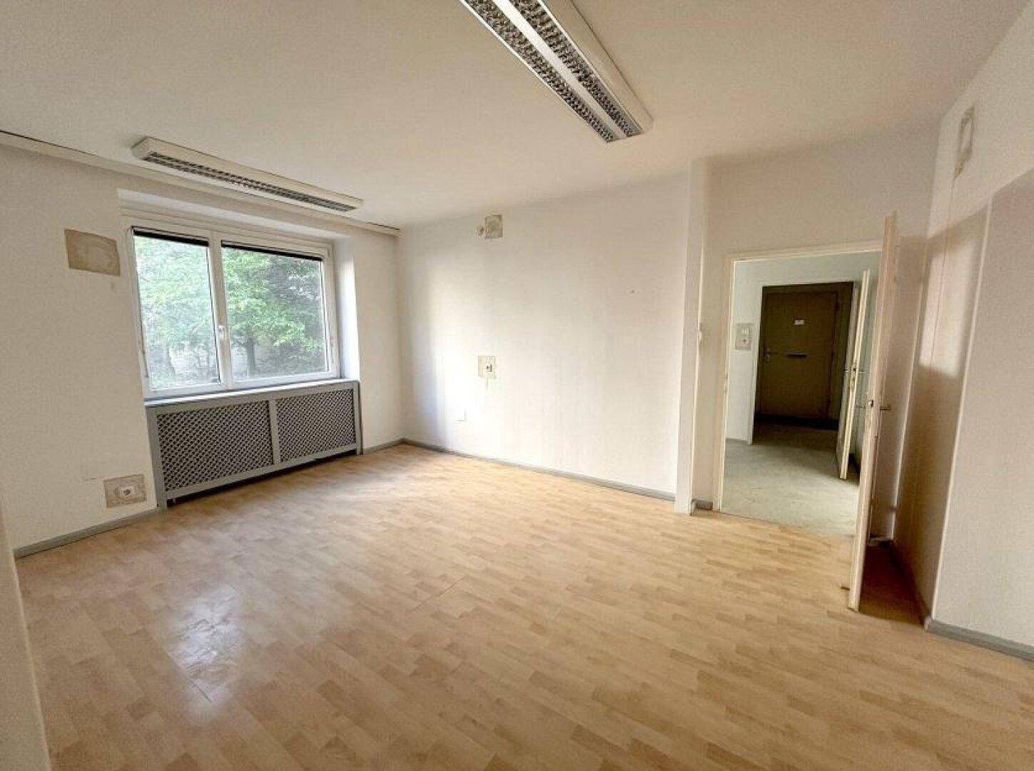 Ein heller Büroraum mit Holzboden und einem Fenster, das den Blick ins Grüne freigibt, sowie einer offenen Tür.