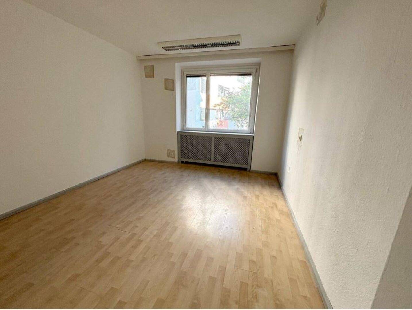Ein kleinerer Büroraum mit hellem Holzboden und einem Fenster, das Tageslicht hereinlässt, sowie einem Heizkörper.
