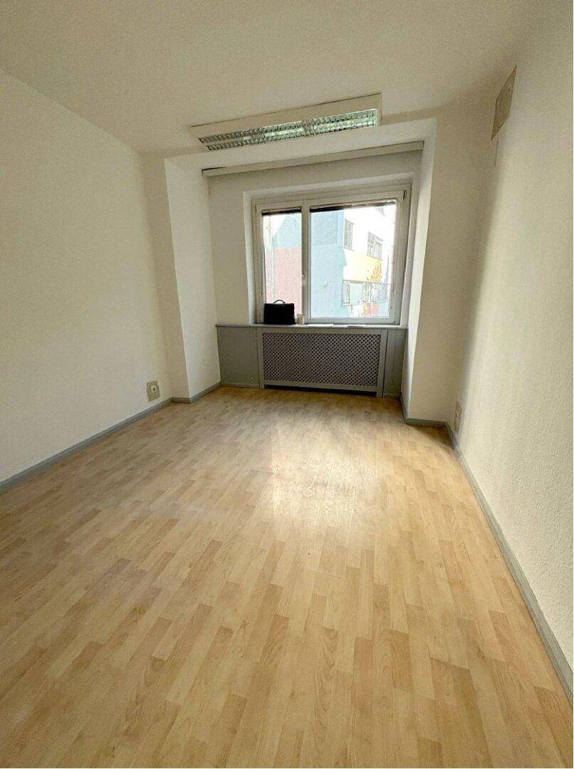 Ein kleinerer Büroraum mit hellem Holzboden, einem Fenster und einem Heizkörper, der eine ruhige Arbeitsumgebung bietet.
