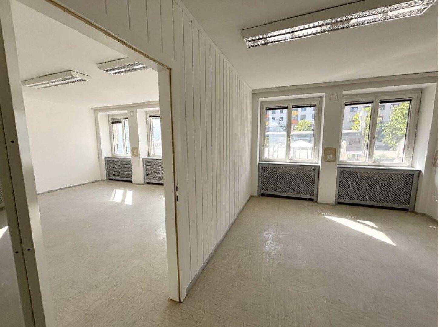 Zwei miteinander verbundene Büroräume mit großen Fenstern, die viel Tageslicht bieten, und einheitlichem Bodenbelag.