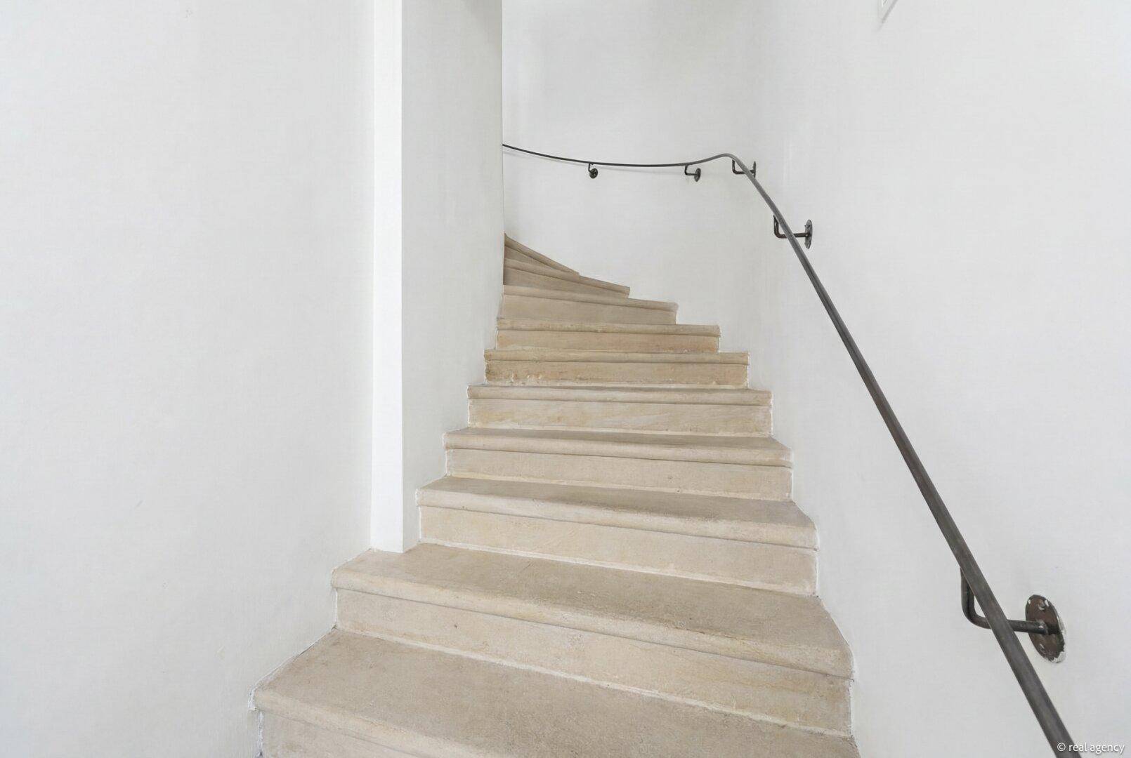 Steintreppe mit schlichtem Metallgeländer in einem hellen Treppenhaus.
