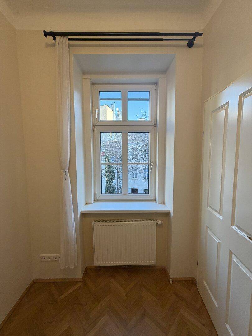 Heller Raum mit Fischgrätparkett, Fenster und Blick auf die umliegenden Gebäude.