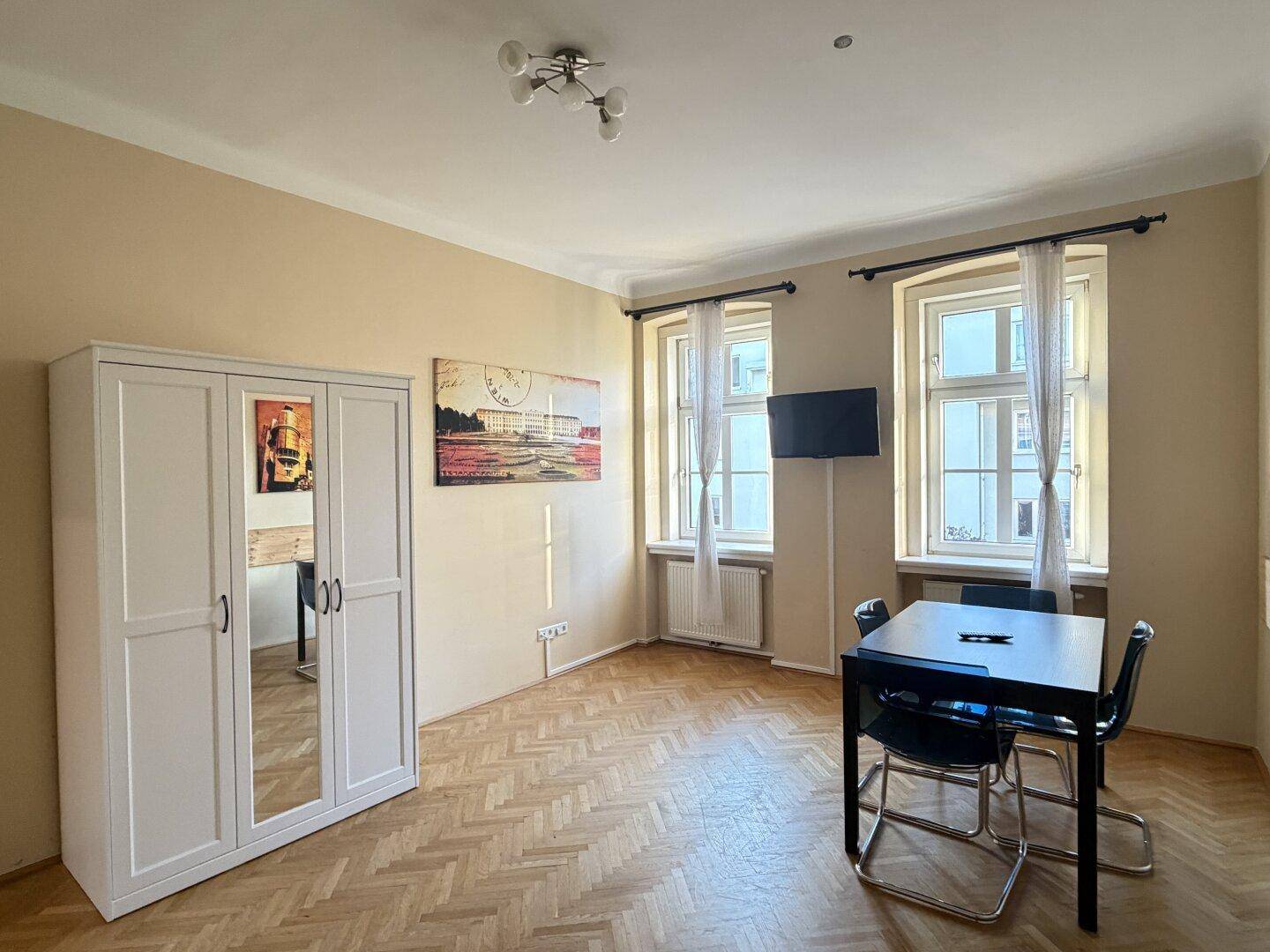 Geräumiges Wohnzimmer mit Fischgrätparkett, weißem Kleiderschrank und viel Tageslicht.