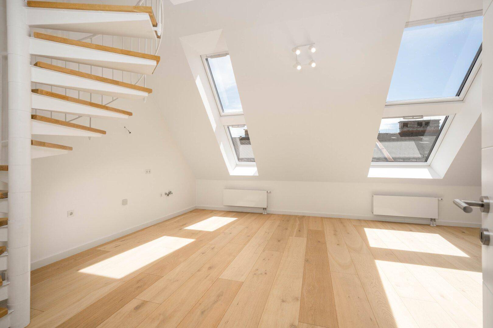 Heller Wohnraum im Dachgeschoss mit Holzfußboden, Wendeltreppe und mehreren Dachfenstern.