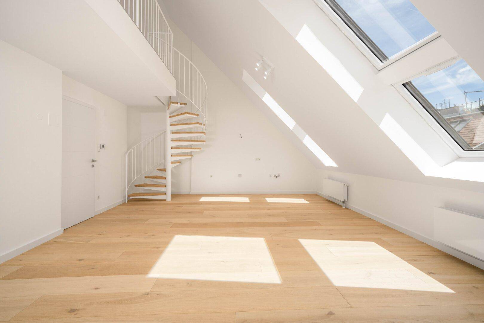 Lichtdurchfluteter Dachgeschossraum mit Holzboden, Wendeltreppe und schrägen Wänden.