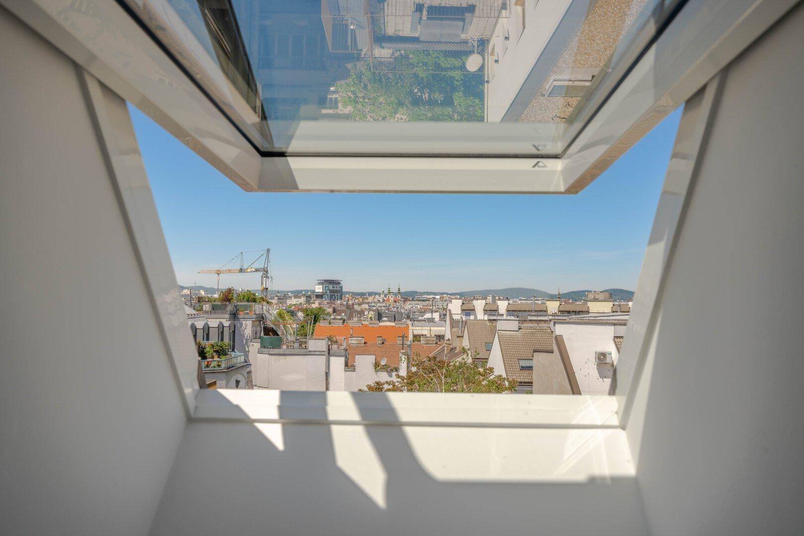 Blick aus einem Dachfenster auf die Stadtlandschaft mit Dächern und blauem Himmel.