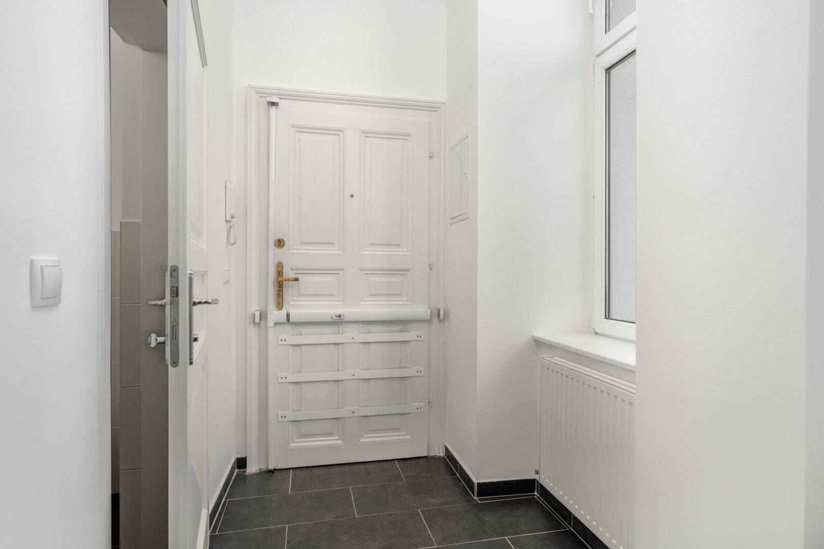 Eingangsbereich mit weißer Tür, Fenster und Heizkörper, ausgestattet mit dunklem Fliesenboden.