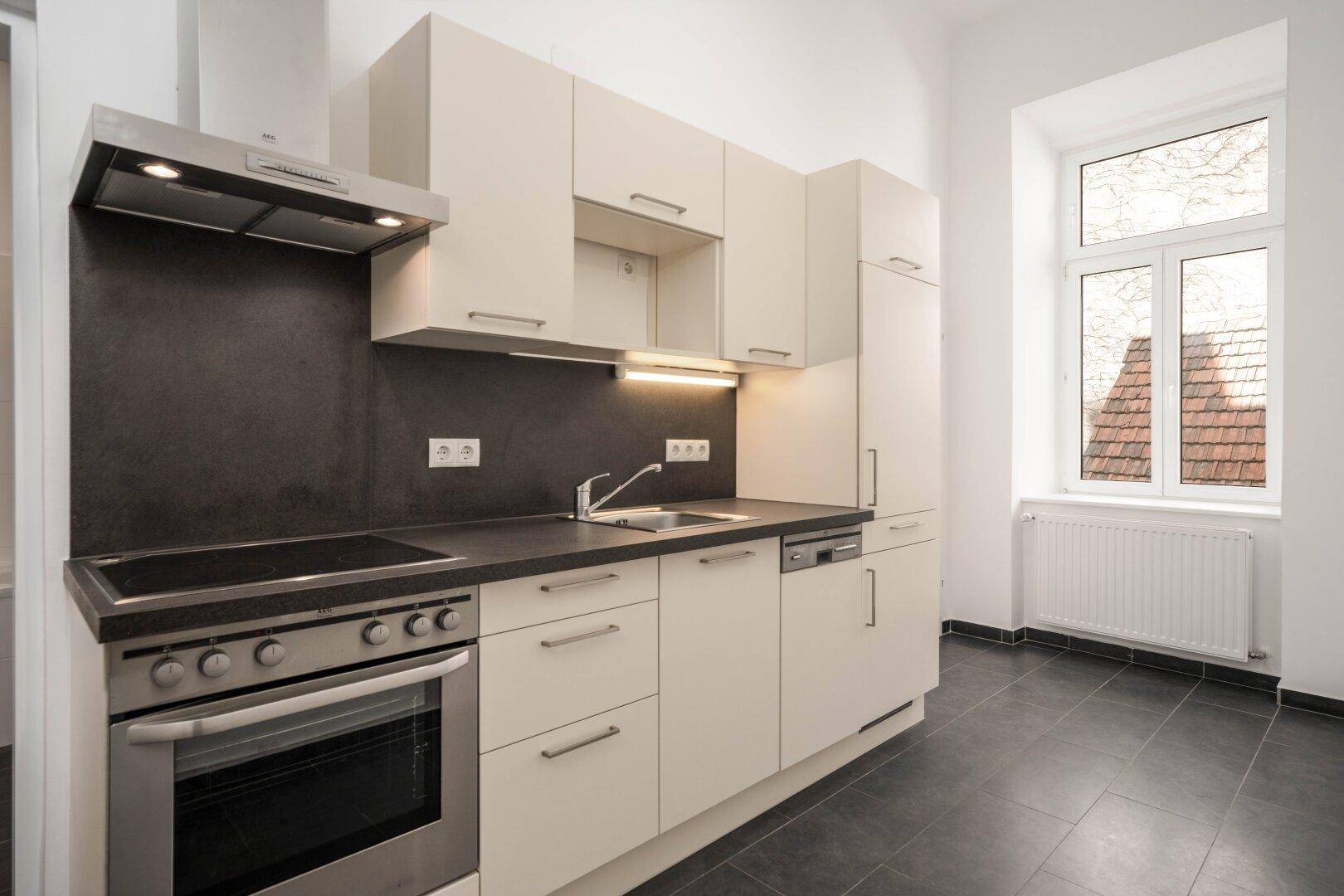Funktionale Einbauküche mit hellen Fronten, dunkler Arbeitsplatte und Fenster mit Blick nach draußen.