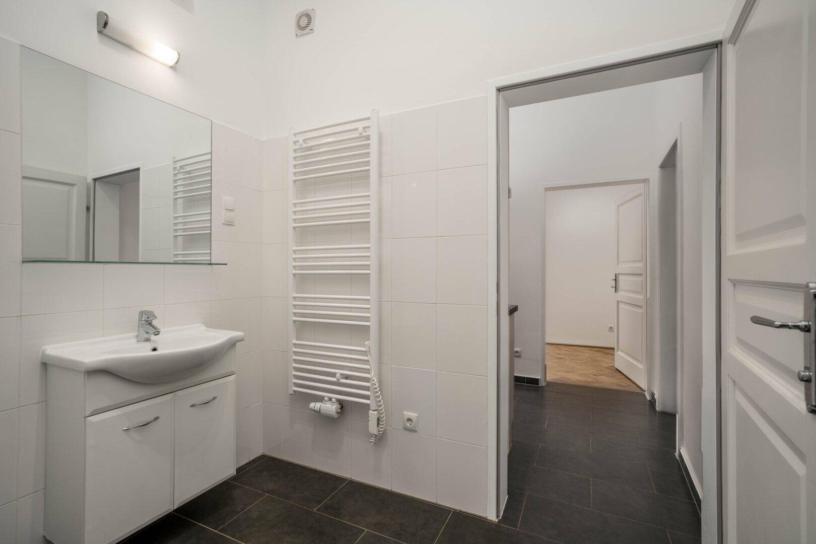 Helles Badezimmer mit Waschbecken, Spiegel und beheiztem Handtuchhalter, gefliestem Boden.