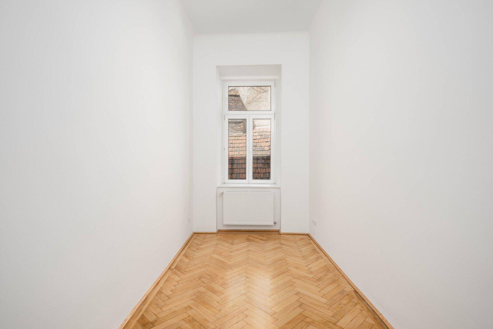 Leerer Raum mit klassischem Fischgrätparkett und einem Fenster, das viel Tageslicht hereinlässt.