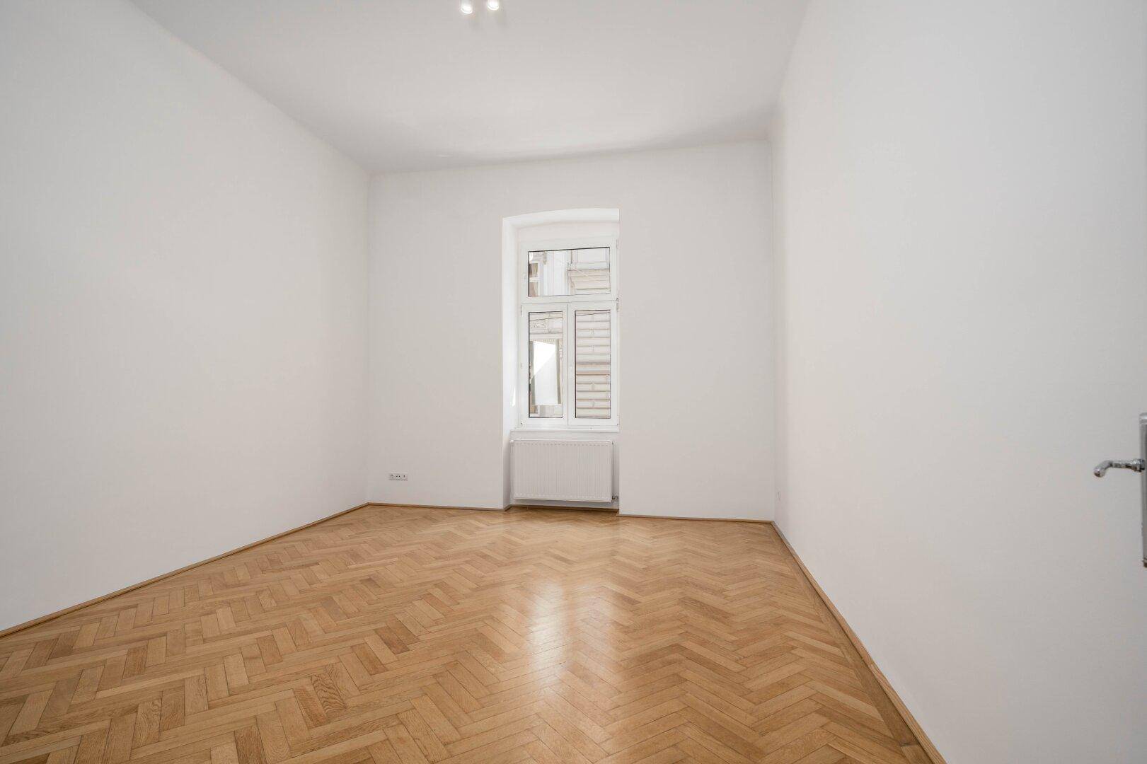 Geräumiger, leerer Raum mit Fischgrätparkett und großem Fenster für natürliche Belichtung.