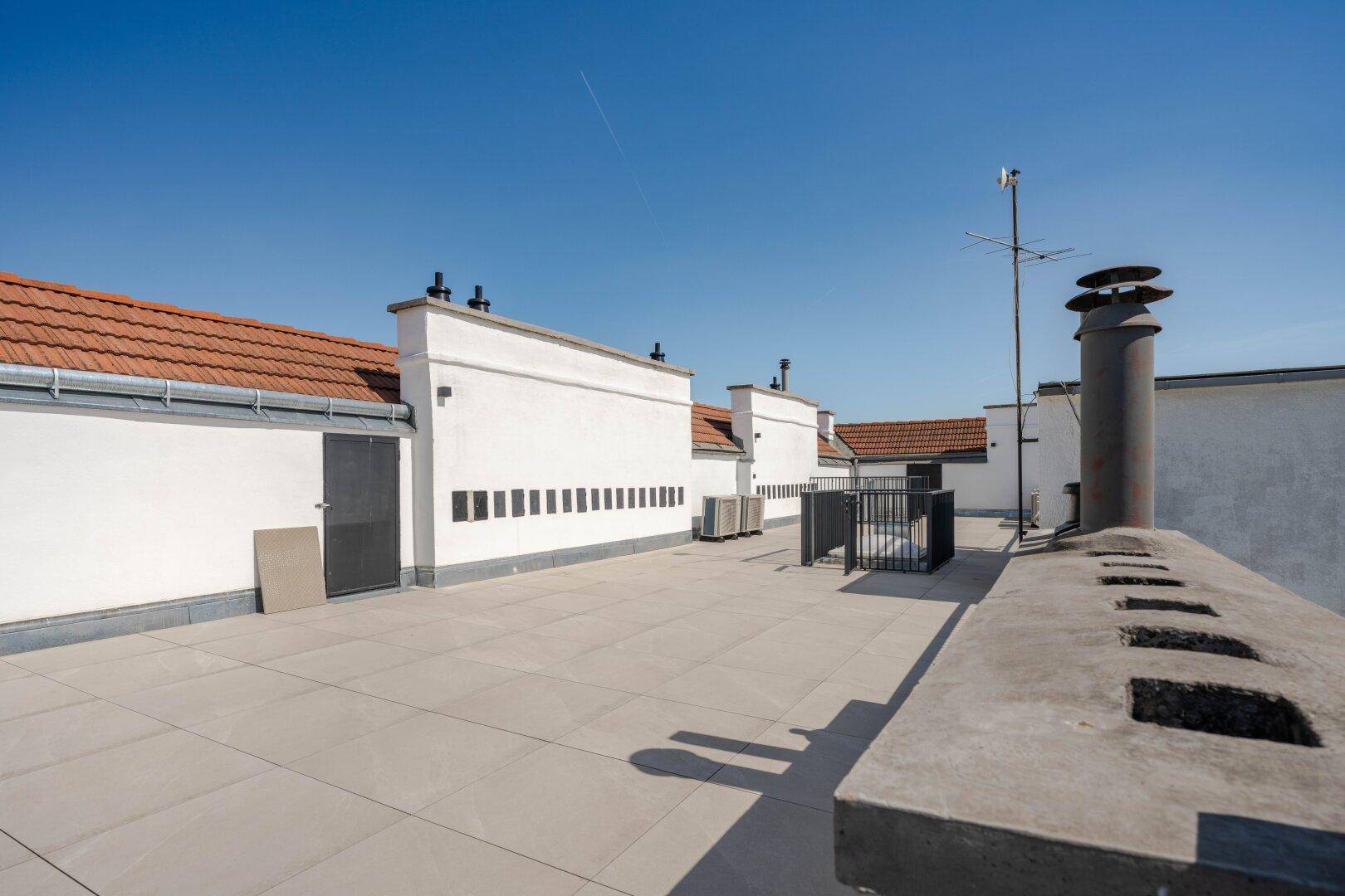 Lange Dachterrasse mit Fliesenboden und mehreren Zugängen, umgeben von Dächern.