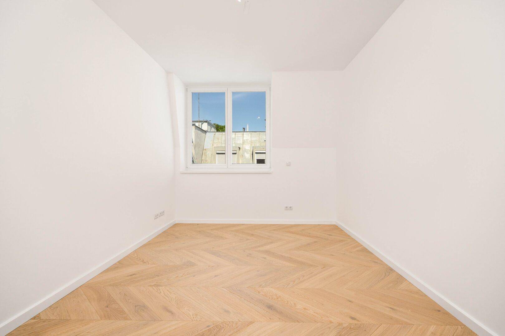 Helles Zimmer mit Fischgrätparkett und einem Fenster mit Blick auf die Umgebung, ideal als Schlafzimmer.