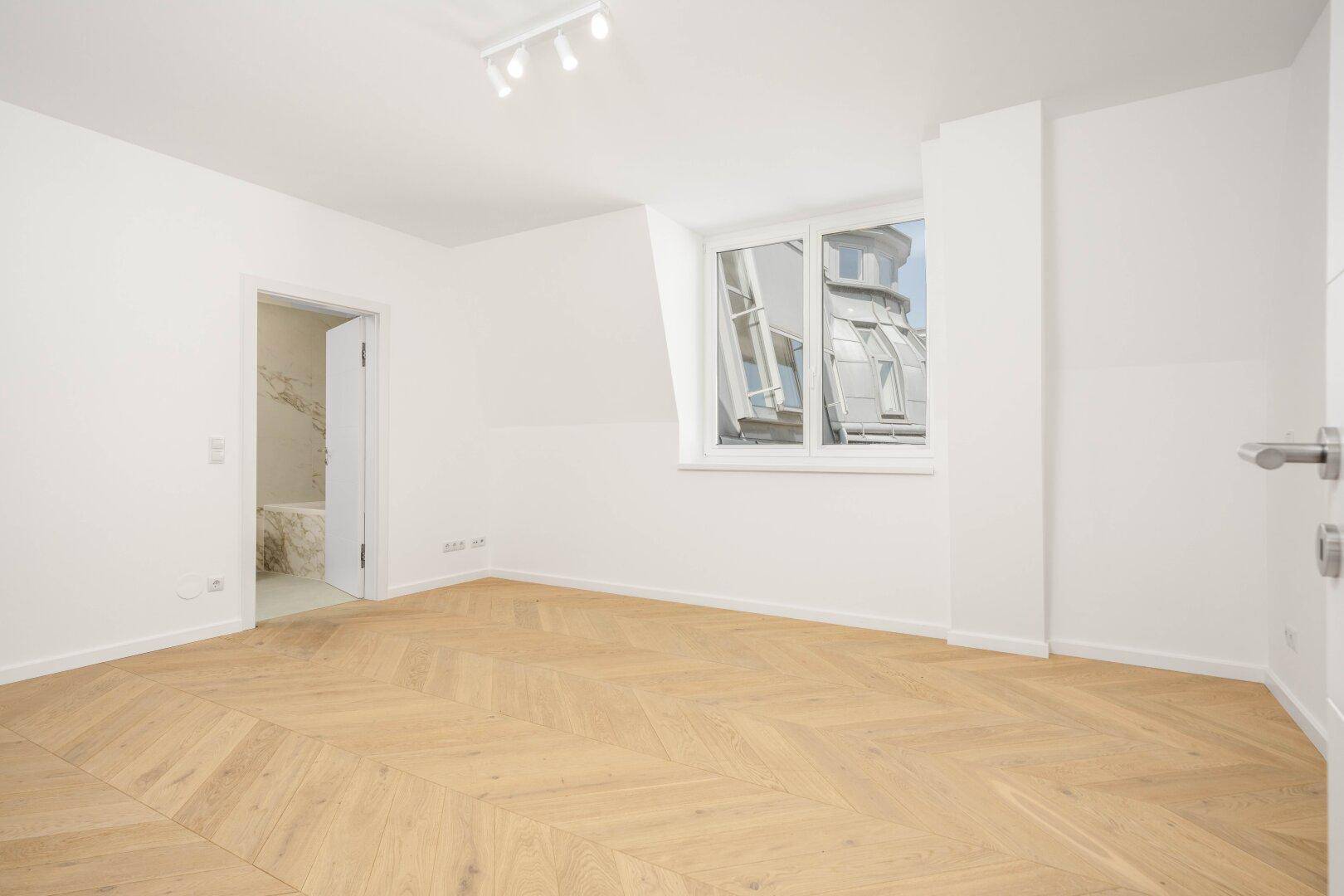 Geräumiges Schlafzimmer mit Fischgrätparkett und direktem Zugang zu einem modernen Badezimmer.