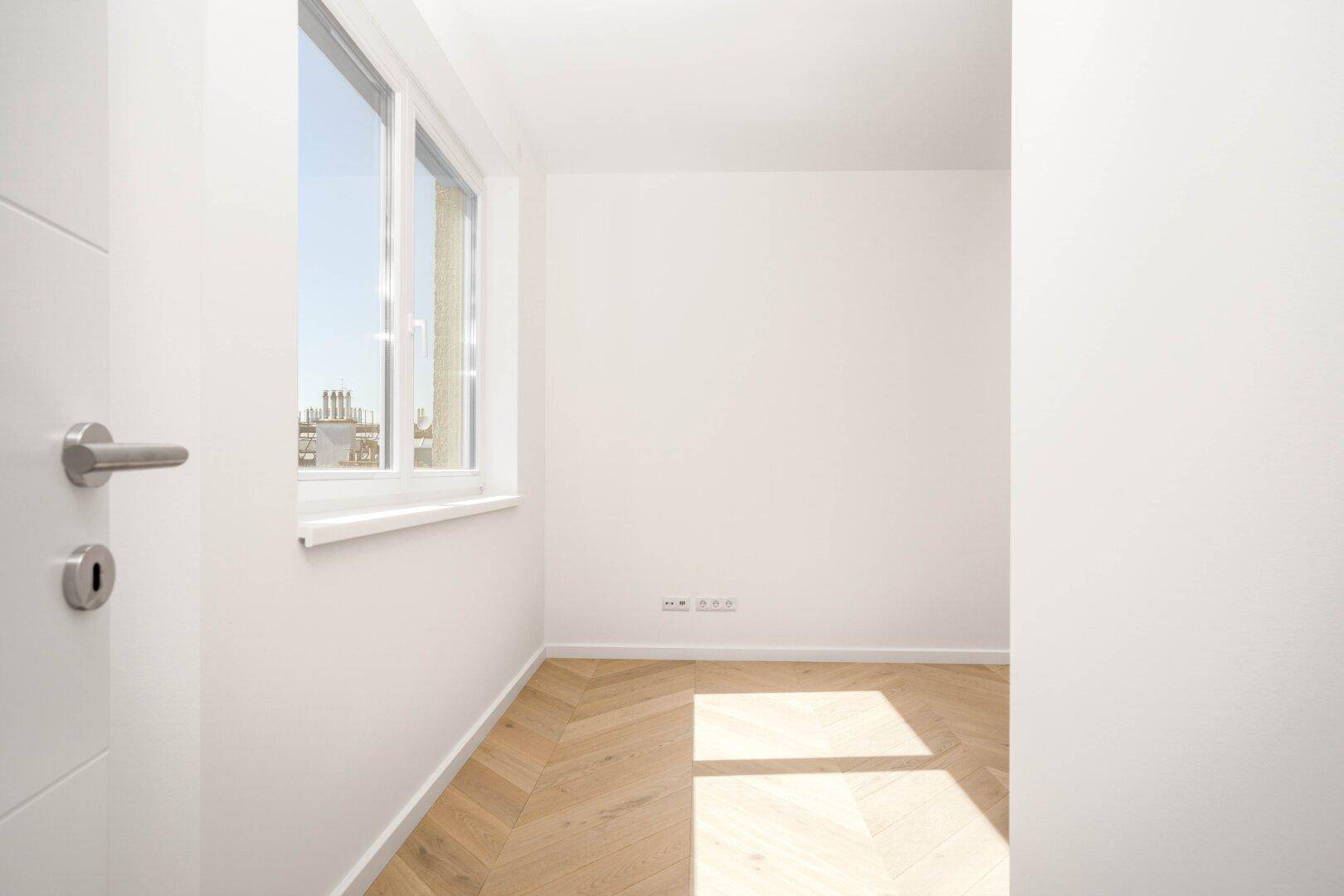 Heller, leerer Raum mit Fischgrätparkettboden und einem großen Fenster, das viel Tageslicht hereinlässt.