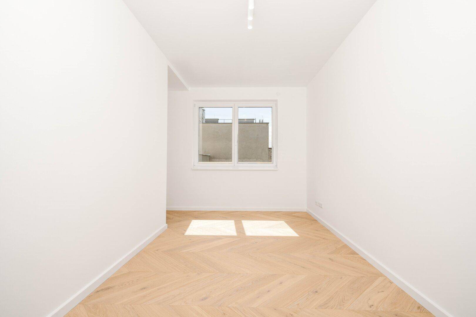Lichtdurchfluteter, leerer Raum mit Fischgrätparkettboden und einem breiten Fenster.