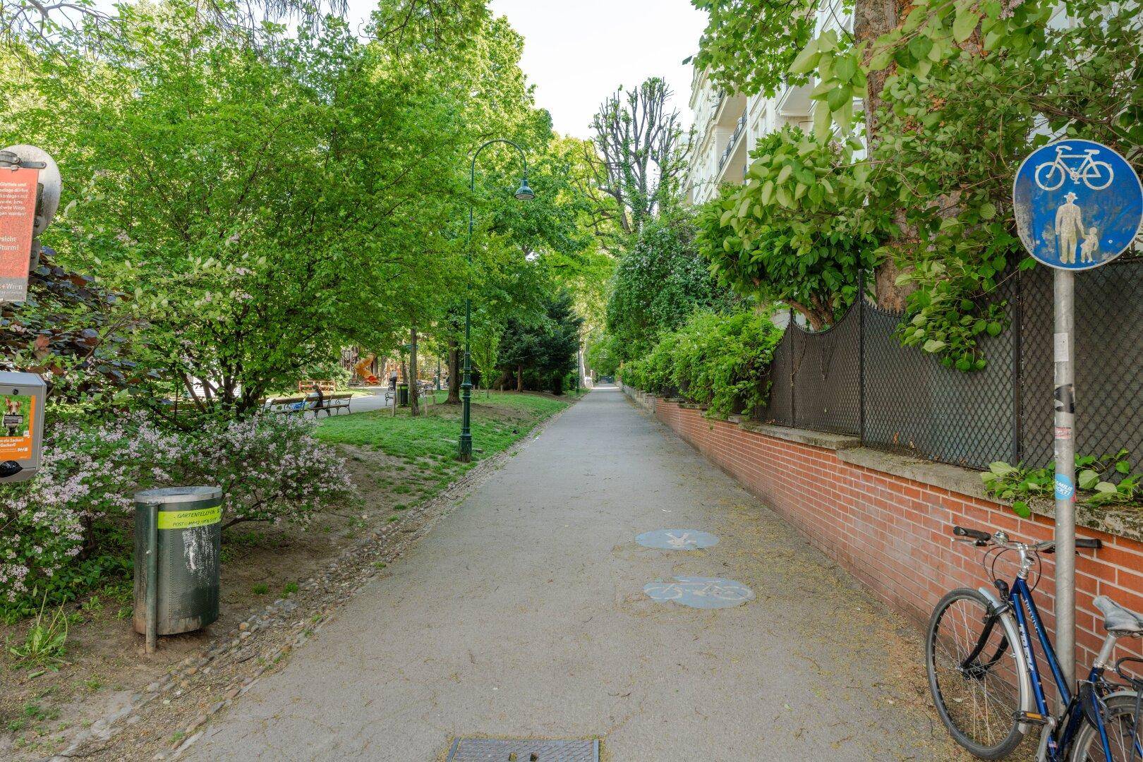 Grüner Parkweg mit Bäumen, einem Fahrrad und einem Hinweisschild für Radfahrer und Fußgänger.