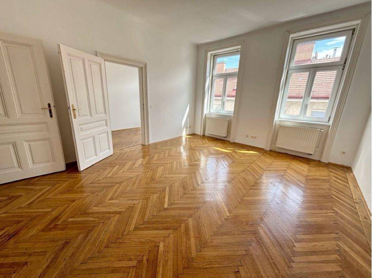 Geräumiger Wohnbereich mit klassischem Fischgrätparkett und hellen Wänden, der durch zwei Fenster belichtet wird.
