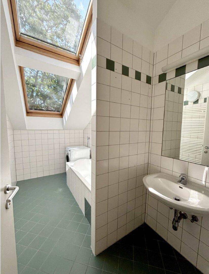 Badezimmer mit weißen Fliesen, grünen Bodenfliesen, Badewanne unter einem Dachfenster und Waschtisch.