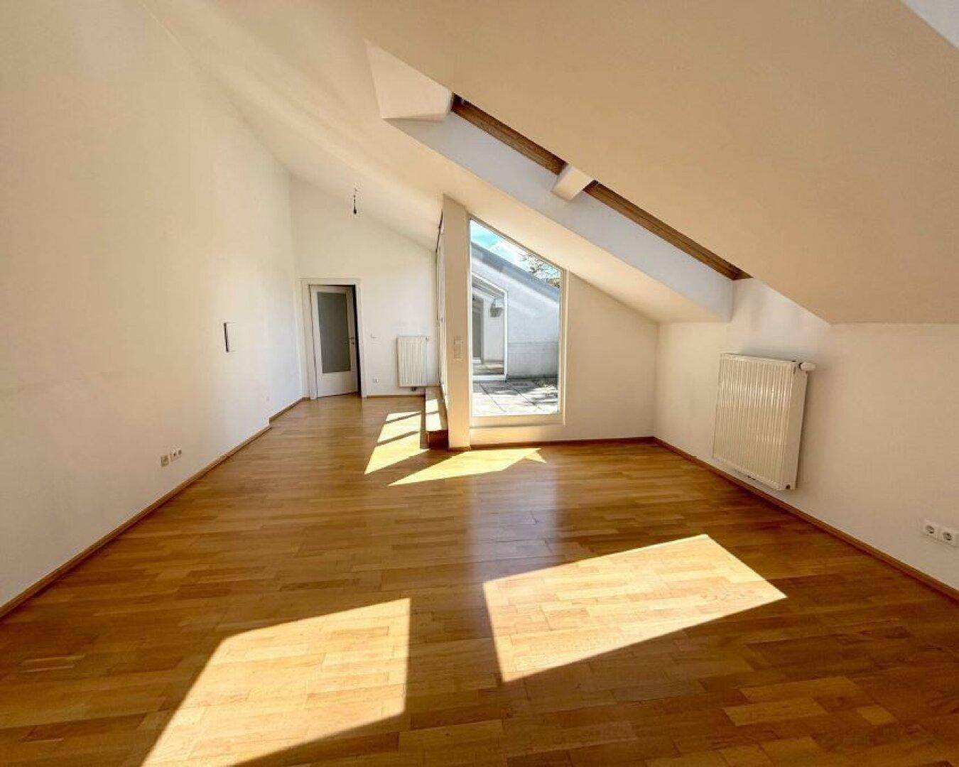 Helles, leeres Zimmer mit Holzparkettboden, Dachschrägen und Zugang zu einer Terrasse.
