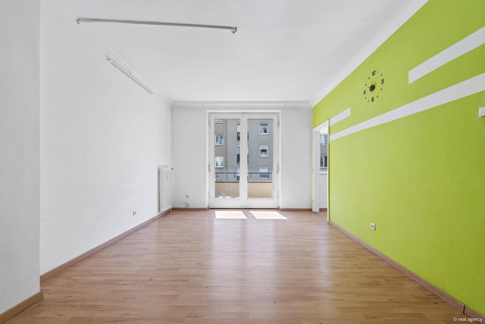 Geräumiges Wohnzimmer mit hellem Holzboden, einer auffälligen grünen Akzentwand und Balkonzugang.