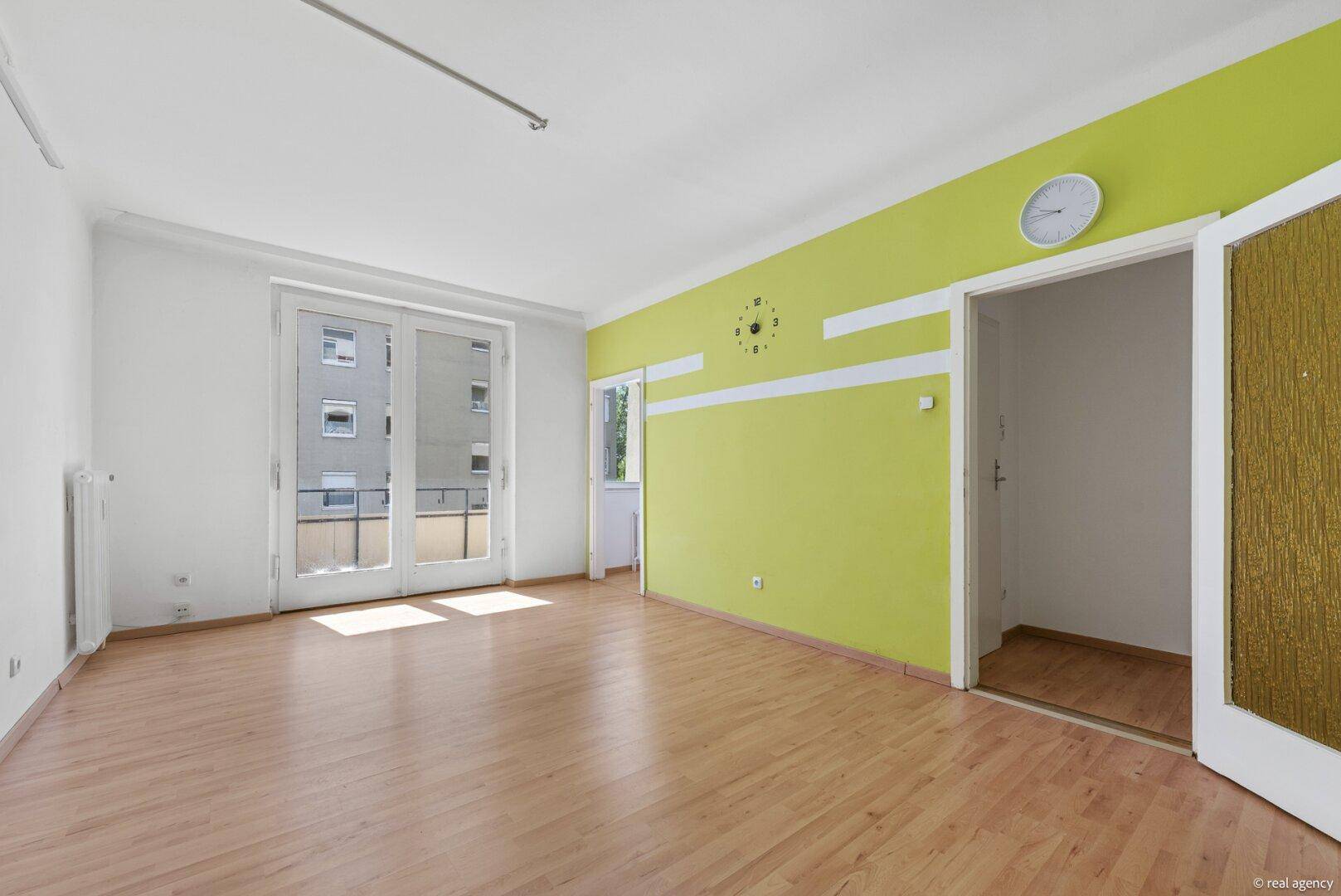 Helles Wohnzimmer mit Holzboden und grüner Wandgestaltung, bietet Zugang zu einem weiteren Raum und Balkon.