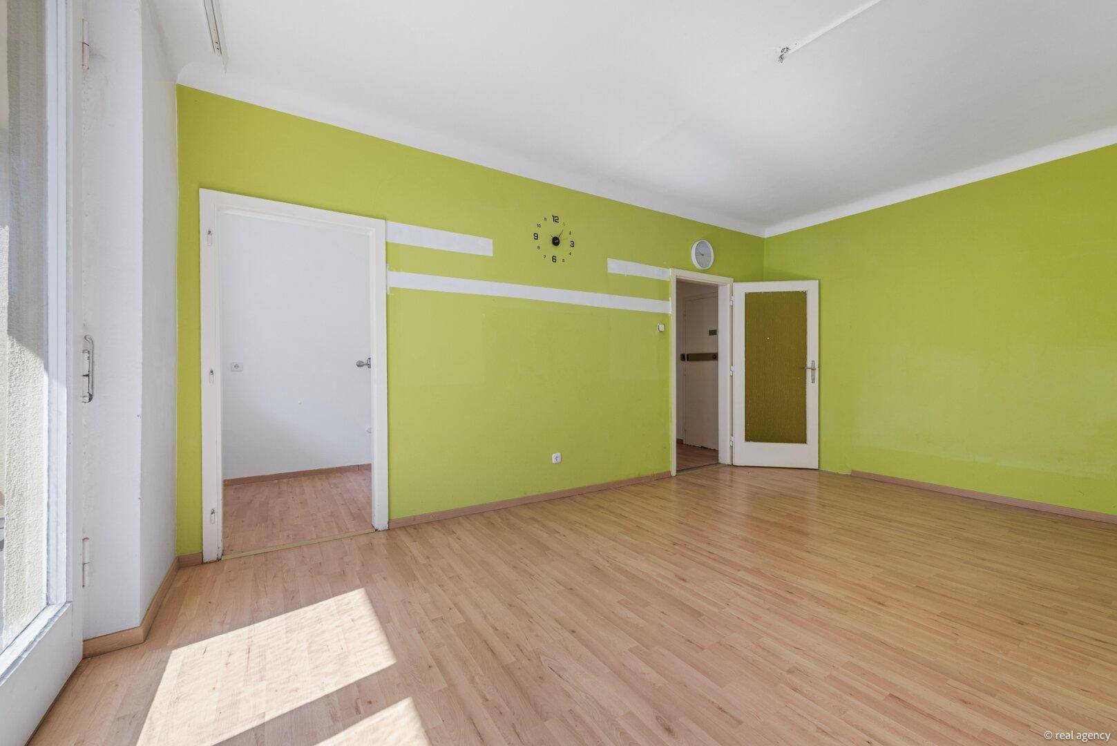 Lichtdurchflutetes Wohnzimmer mit Holzboden und grüner Akzentwand, mit zwei Türen zu angrenzenden Räumen.