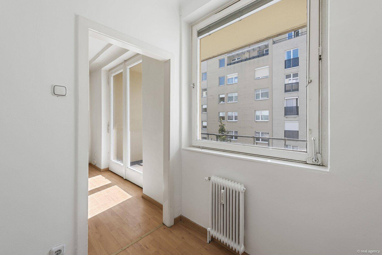 Heller Flurbereich mit Fenster und Heizkörper, der zu einem weiteren Raum mit Balkonzugang führt.