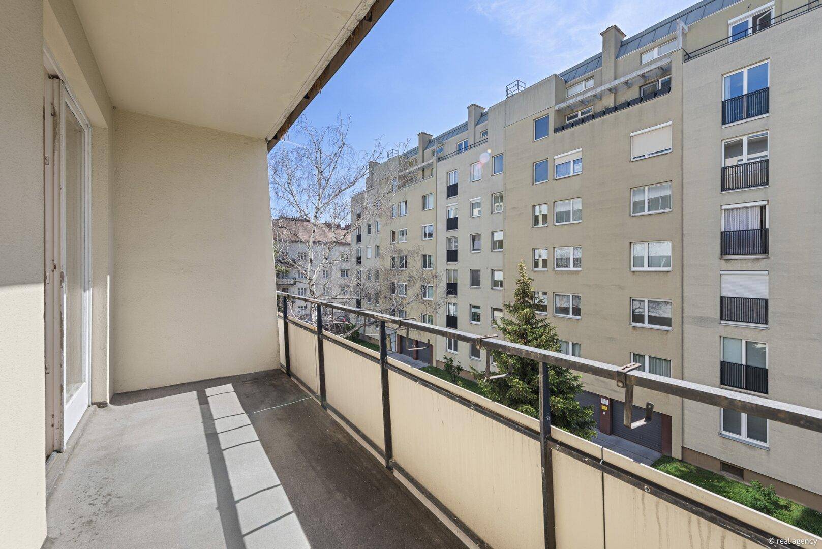 Geräumiger Balkon mit Blick auf die gegenüberliegende Wohnbebauung und einige Bäume im Frühling.