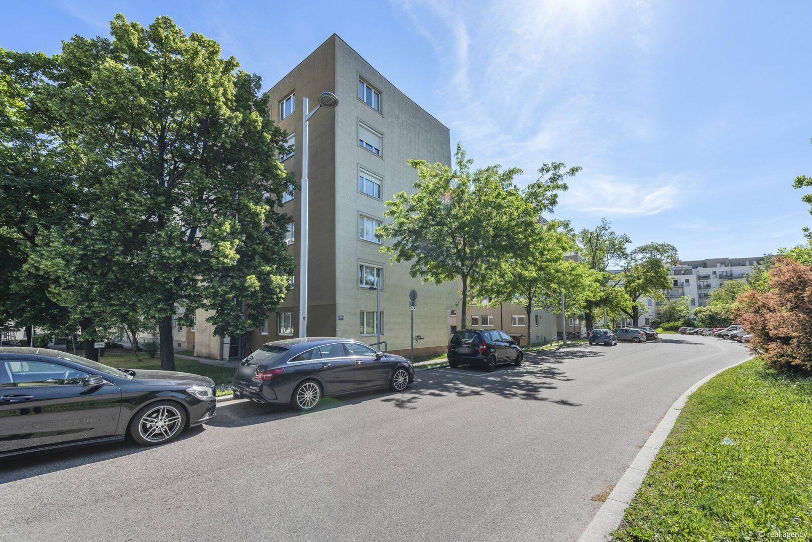 Mehrfamilienhaus mit schlichter Fassade, umgeben von Bäumen und Parkplätzen an der Straße.