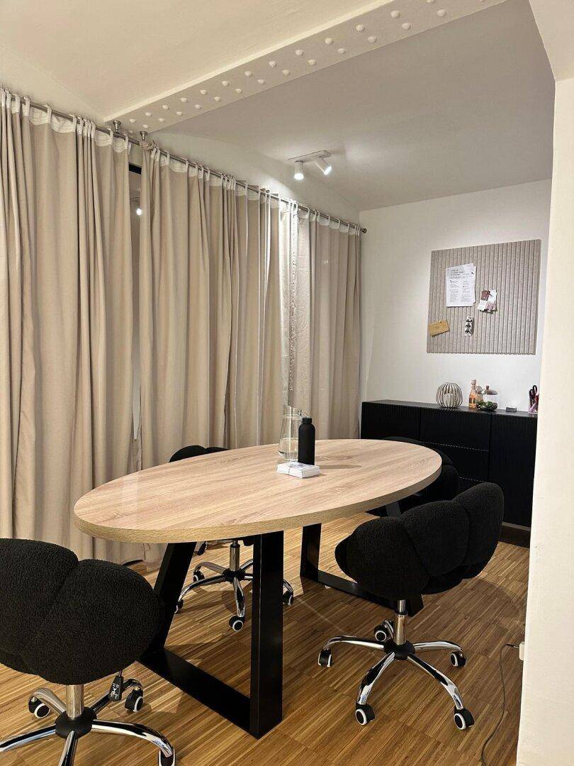 Gemütlicher Büroraum mit ovalem Holztisch und schwarzen Stühlen, abgetrennt durch helle Vorhänge für Privatsphäre.