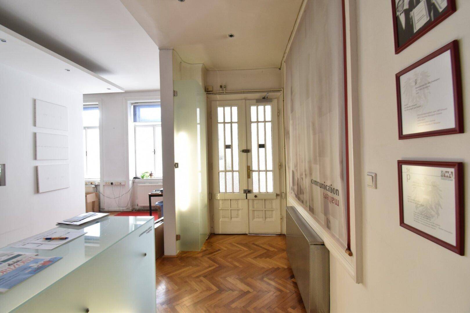 Heller Flur mit Fischgrätparkett, einer modernen Rezeptionstheke und gerahmten Dokumenten an der Wand.