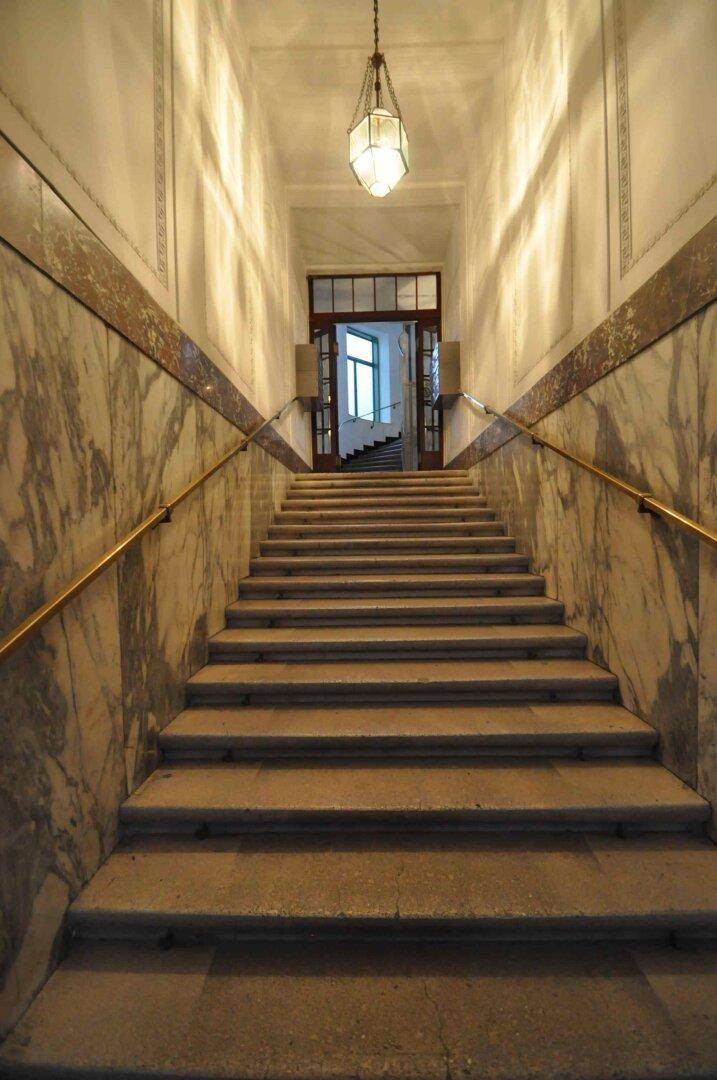 Repräsentatives Treppenhaus mit Marmorverkleidung, breiten Stufen und einem eleganten Kronleuchter.