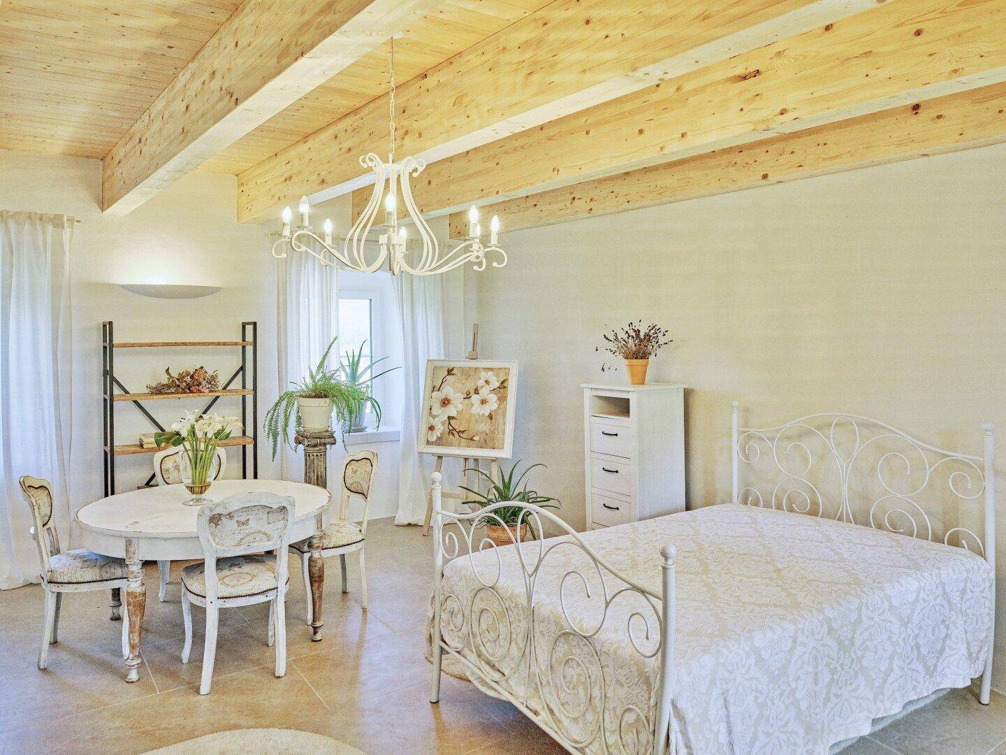 Geräumiges Schlafzimmer mit Holzbalkendecke, weißem Schmiedeeisenbett und viel Tageslicht.