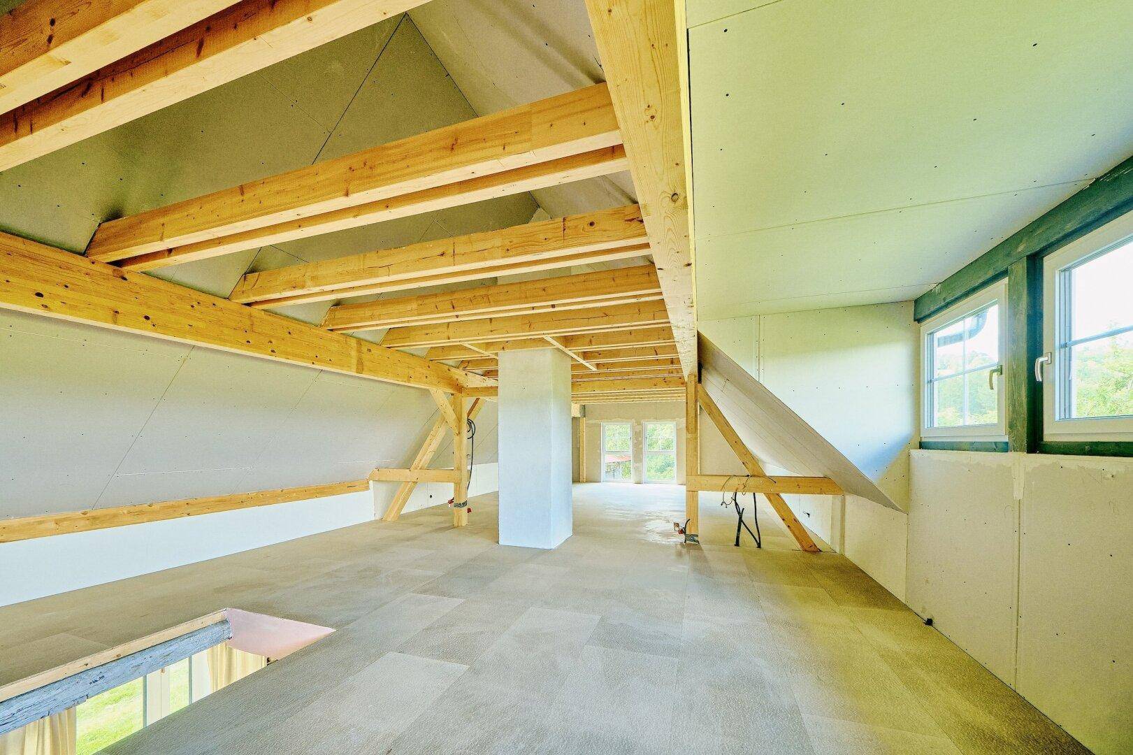 Weitläufiger Dachboden im Rohbau mit sichtbarer Dachschräge und mehreren Fenstern für Tageslicht.