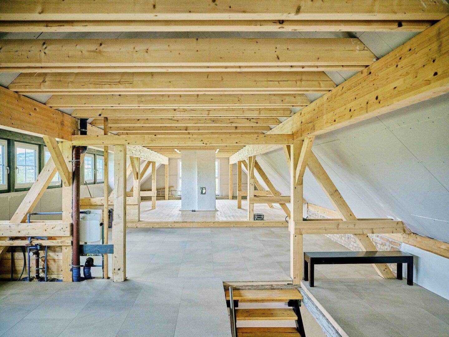 Weiterer Blick in den unfertigen Dachboden mit Holzbalken, Treppenzugang und sichtbaren Installationen.