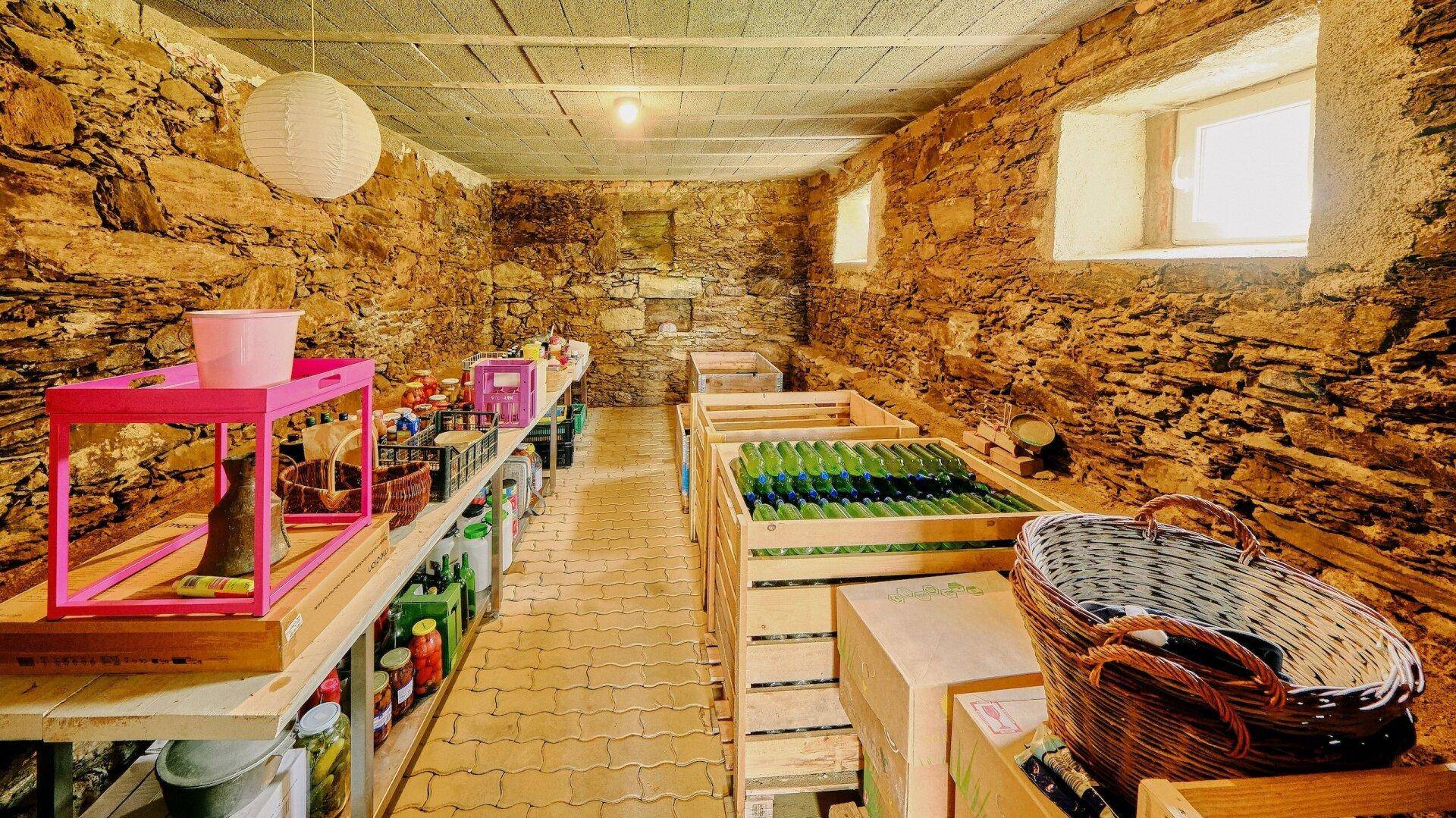 Rustikaler Kellerraum mit Natursteinwänden, gefüllt mit Weinflaschen und Vorräten in Holzkisten.