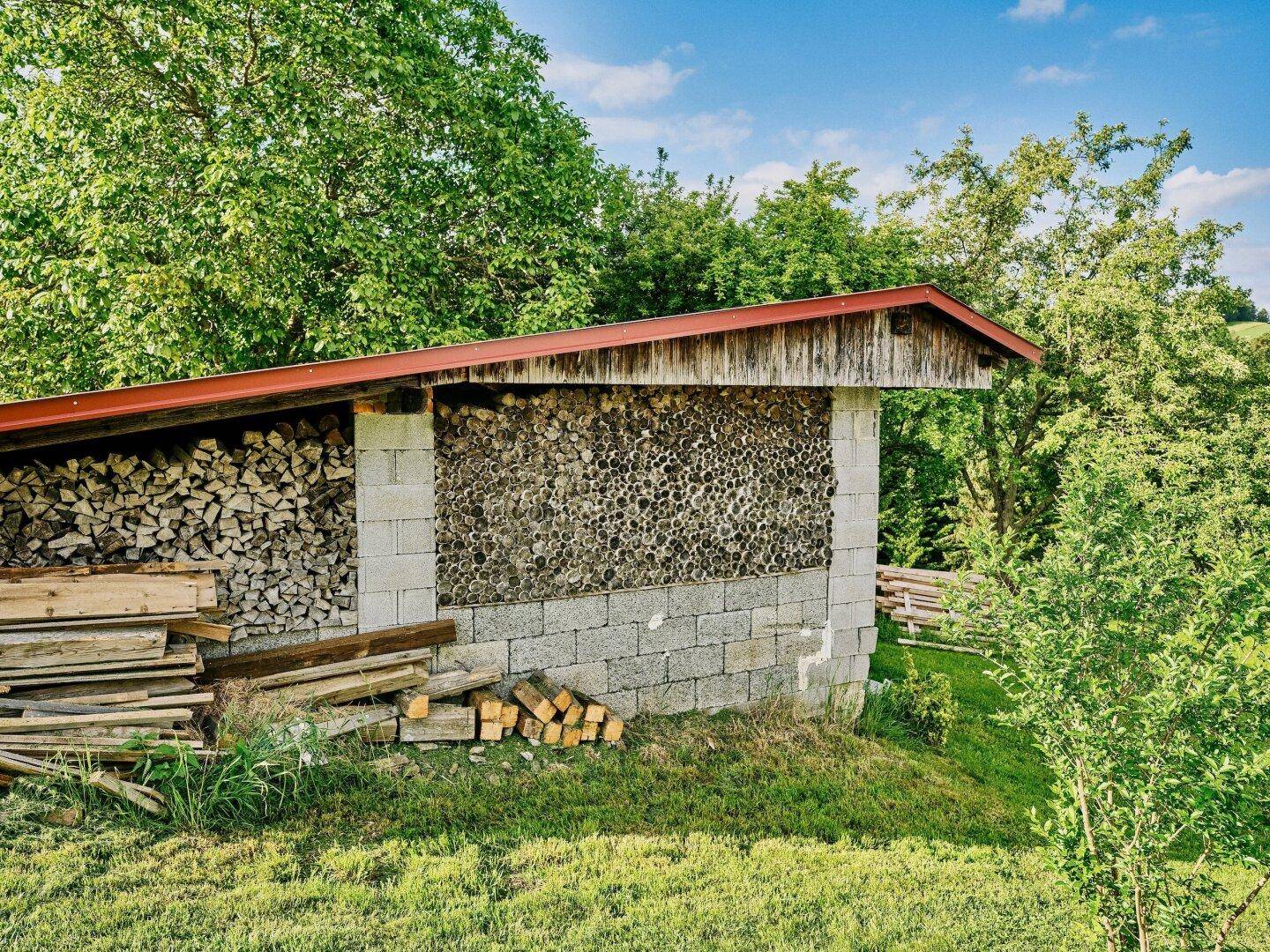 Rustikaler Holzschuppen mit gestapeltem Brennholz, umgeben von grüner Natur und Bäumen.