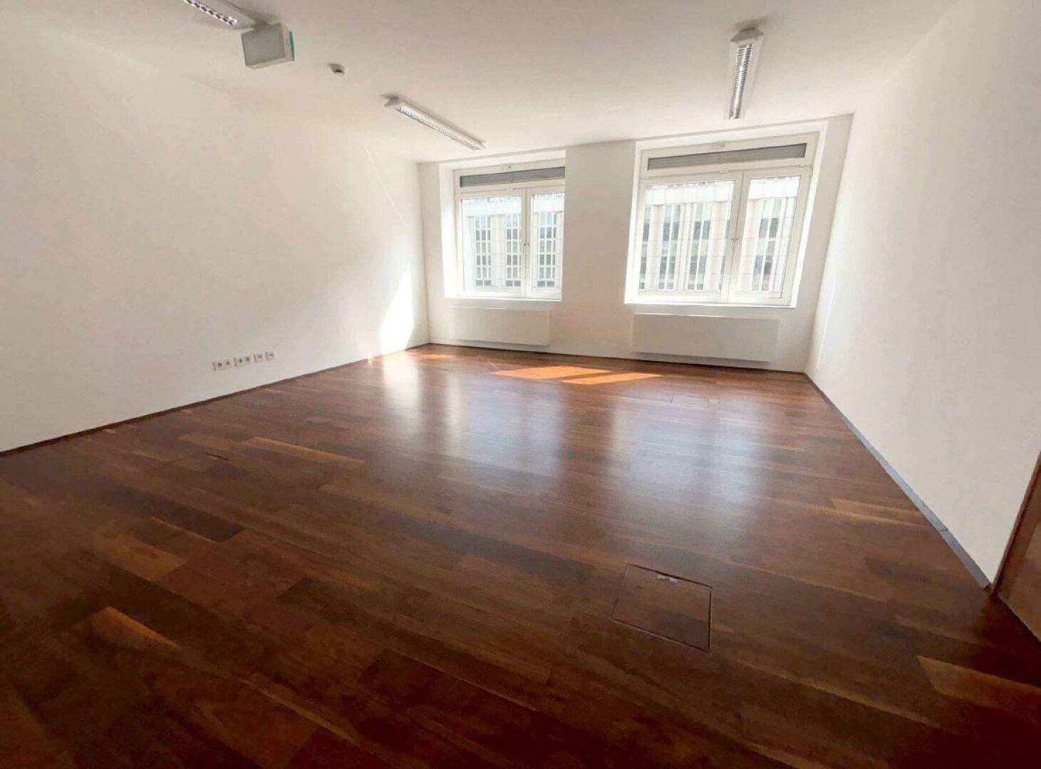 Heller, leerer Büroraum mit glänzendem Holzboden und zwei großen Fenstern, die viel Tageslicht hereinlassen.