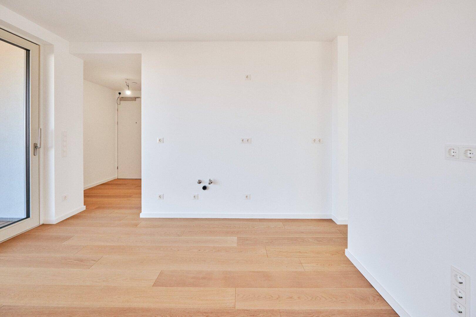 Leerer Küchenbereich mit Anschlüssen für Geräte, Holzboden und Zugang zum Balkon in Neubauqualität.