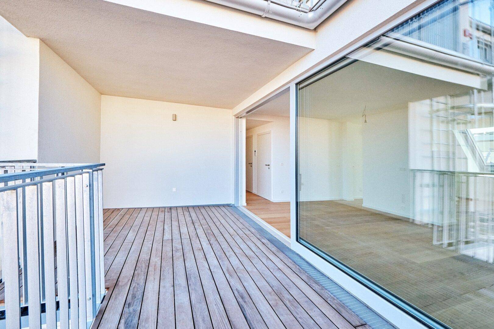 Geräumiger Balkon mit Holzbelag und Glasfront, verbunden mit dem hellen Innenbereich.