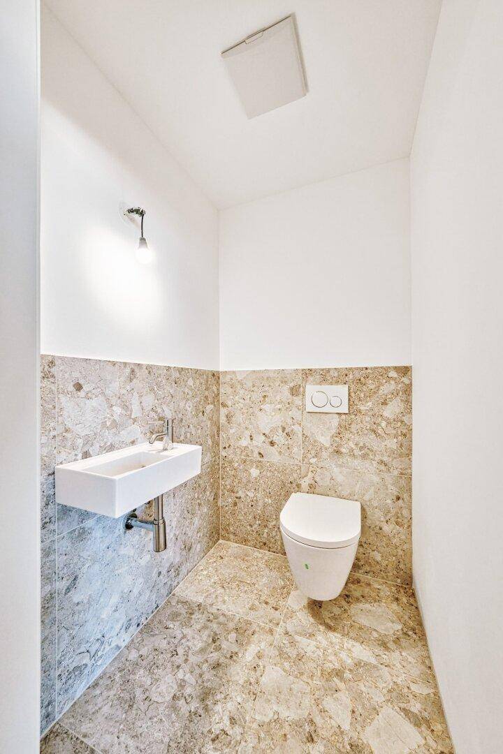 Separates Gäste-WC mit wandhängender Toilette, kleinem Waschbecken und Steinfliesen in Neubauqualität.