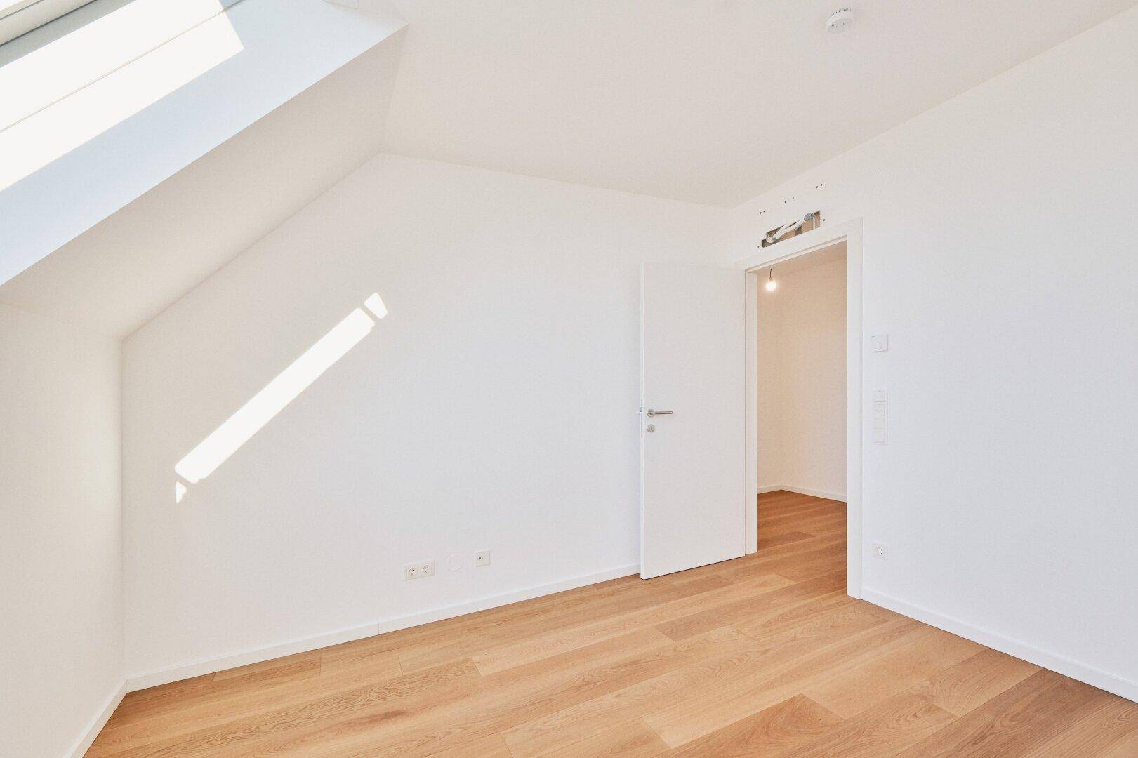 Leeres Dachgeschosszimmer mit Holzboden, schrägen Wänden und viel Tageslicht in Neubauqualität.